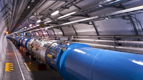 A long curving tunnel at CERN lined with the blue cylindrical superconducting magnets of the Large Hadron Collider, with cable trays and lights along the wall.