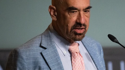 David Spergel, a bald man with a grey beard in a light blue checked jacket and pink tie, speaking beside a microphone.