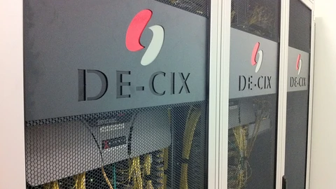 Grey mesh cabinet doors marked DE-CIX, with yellow fibre visible behind the mesh. No people appear.