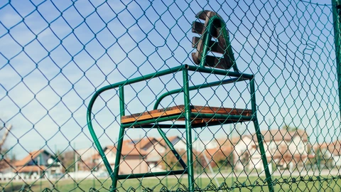 An empty green umpire chair behind a fence overlooking a red clay tennis court. No people appear.