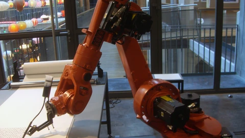An orange KUKA industrial robot arm writing on a surface in a workshop. No people appear.