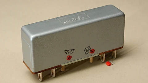 A grey, hand-sized Soviet-era DP-29 latching relay with screw terminals on a beige background, next to a tiny red component for scale.