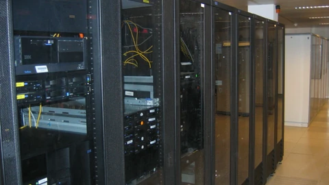 Aisle between tall server racks in a data center with blue cabinet doors and overhead cable trays. No people appear.