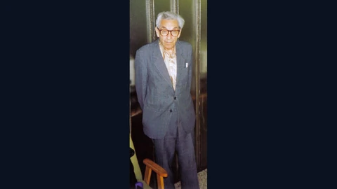 An archival photo of the mathematician Paul Erdős, an elderly man in glasses and a grey suit, standing in a doorway at a student seminar in Budapest.