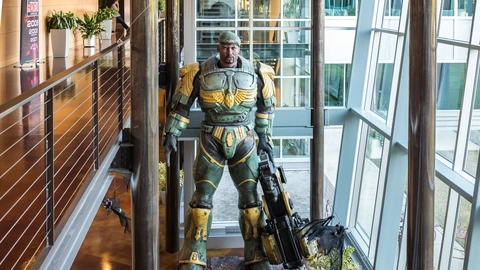 Inside a glass-walled atrium with wooden floors and exposed ducts, a life-size statue of an armoured soldier holding a large rifle stands between tall window frames, with plants and a railing in front.