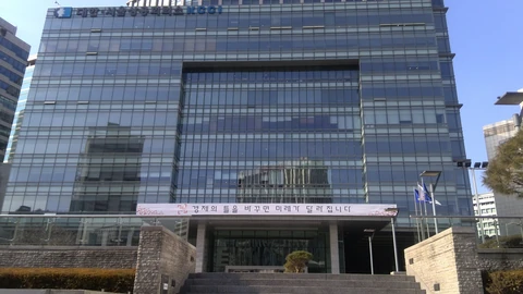 A tall glass office tower with a wide stepped entrance and flagpoles, the Korea Chamber of Commerce and Industry building in Seoul, under a blue sky.