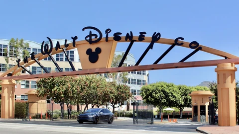 The arched Alameda Avenue gate of the Walt Disney Studios in Burbank, with ‘The Walt Disney Co.’ lettering and a Mickey Mouse emblem above a driveway, office buildings and trees behind it.