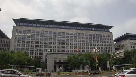 A long, flat-roofed grey ministry building with rows of windows and a red flag above the entrance, seen behind trees, a fenced avenue and passing cars under an overcast sky.