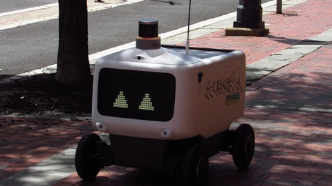A white Avride delivery robot with scrawled marks on its side rolling along a brick sidewalk in Philadelphia, past a tree and a black post.