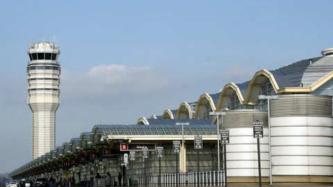 The terminal entrance at Ronald Reagan Washington National Airport with its glass and steel facade and the airport air traffic control tower rising behind it under a blue sky.