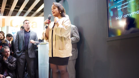 Jaya Baloo speaking into a handheld microphone at a trade show stand in 2014, wearing a cream jacket, with listeners standing behind her and a presentation screen to the right.