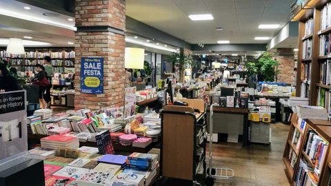 Rows of tables piled with books and tall wooden shelves inside a large Kyobo bookstore, with a brick pillar and a few distant shoppers.