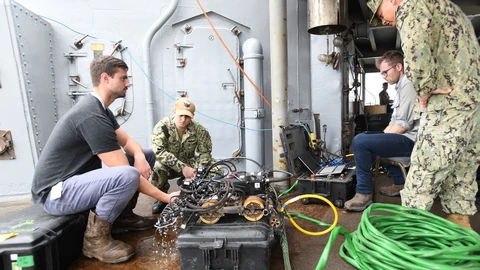 A Gecko Robotics engineer and a Navy sailor crouch beside a tracked inspection robot covered in cables on a wet steel deck, while another engineer watches a rugged laptop and a sailor in camouflage stands nearby.