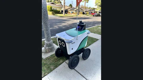 A white and green Serve Robotics delivery robot with two round headlight eyes and a small screen that reads Sula is on duty, parked on a grass-lined residential sidewalk.