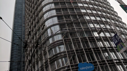 Salesforce Tower seen from the street on an overcast day, with the Salesforce cloud mark on the glass podium and tram wires across the frame. No people appear.