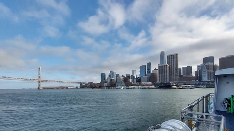 San Francisco’s South of Market skyline across the bay from a ferry, with the Bay Bridge span at left, Salesforce Tower among the towers, and a white ferry railing at right. No people appear.