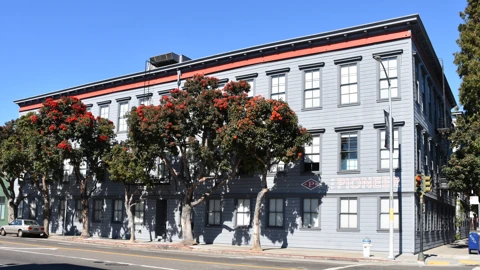 Corner of a grey clapboard office building with a red cornice and a PIONEERER sign, flowering street trees, a silver sedan parked at left, and a clear sky. No people appear.