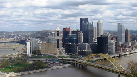 Pittsburgh skyline under a blue sky with bridges in the foreground. No identifiable people appear.