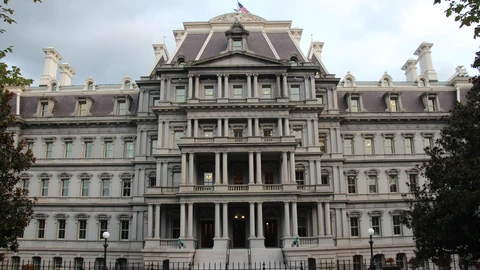 The grey south facade of the Eisenhower Executive Office Building behind a black iron fence, with a flag on the roof and empty steps. No people appear.