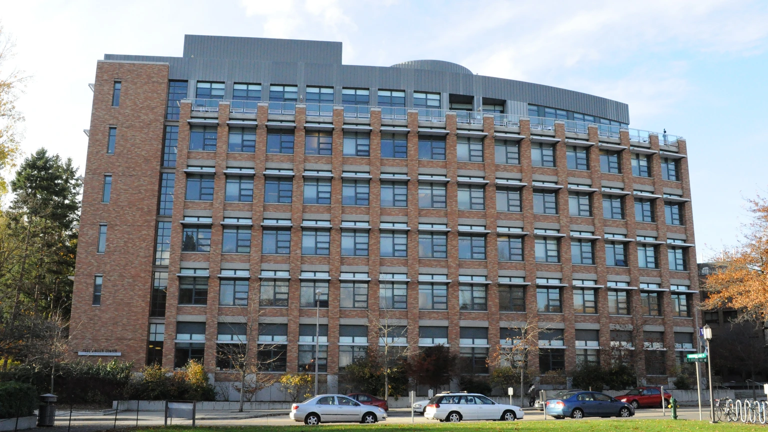 A seven-storey brick university building with regular window bays, a roof terrace and three parked cars on a lawn in front.