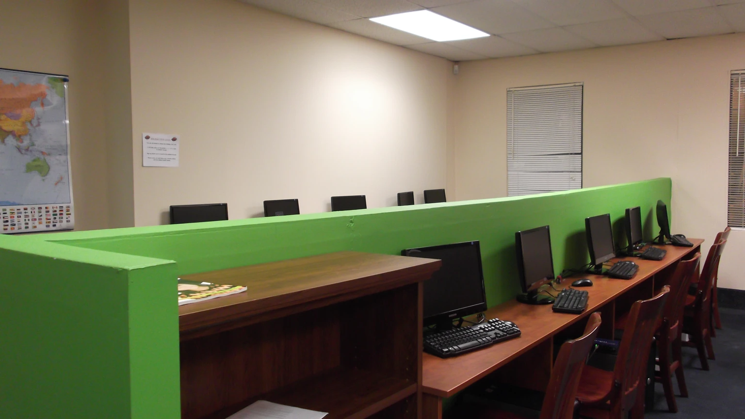 A row of desktop computers on a wooden desk behind a bright green partition in an empty college computer lab.