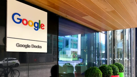 A white Google logo sign reading Google Docklands beside a glass office entrance with potted shrubs.