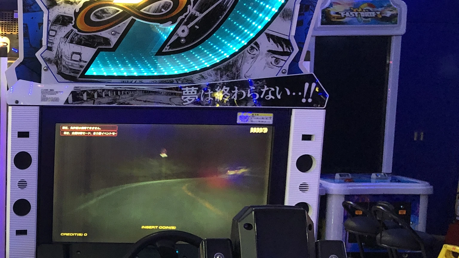 Close view of an empty Initial D Arcade Stage cabinet, showing the wheel, screen and illuminated marquee.