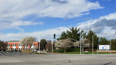 The entrance road to Facebook’s Menlo Park campus, with a low office building, blossoming trees and a white sign showing a thumbs-up icon and 1 Hacker Way.