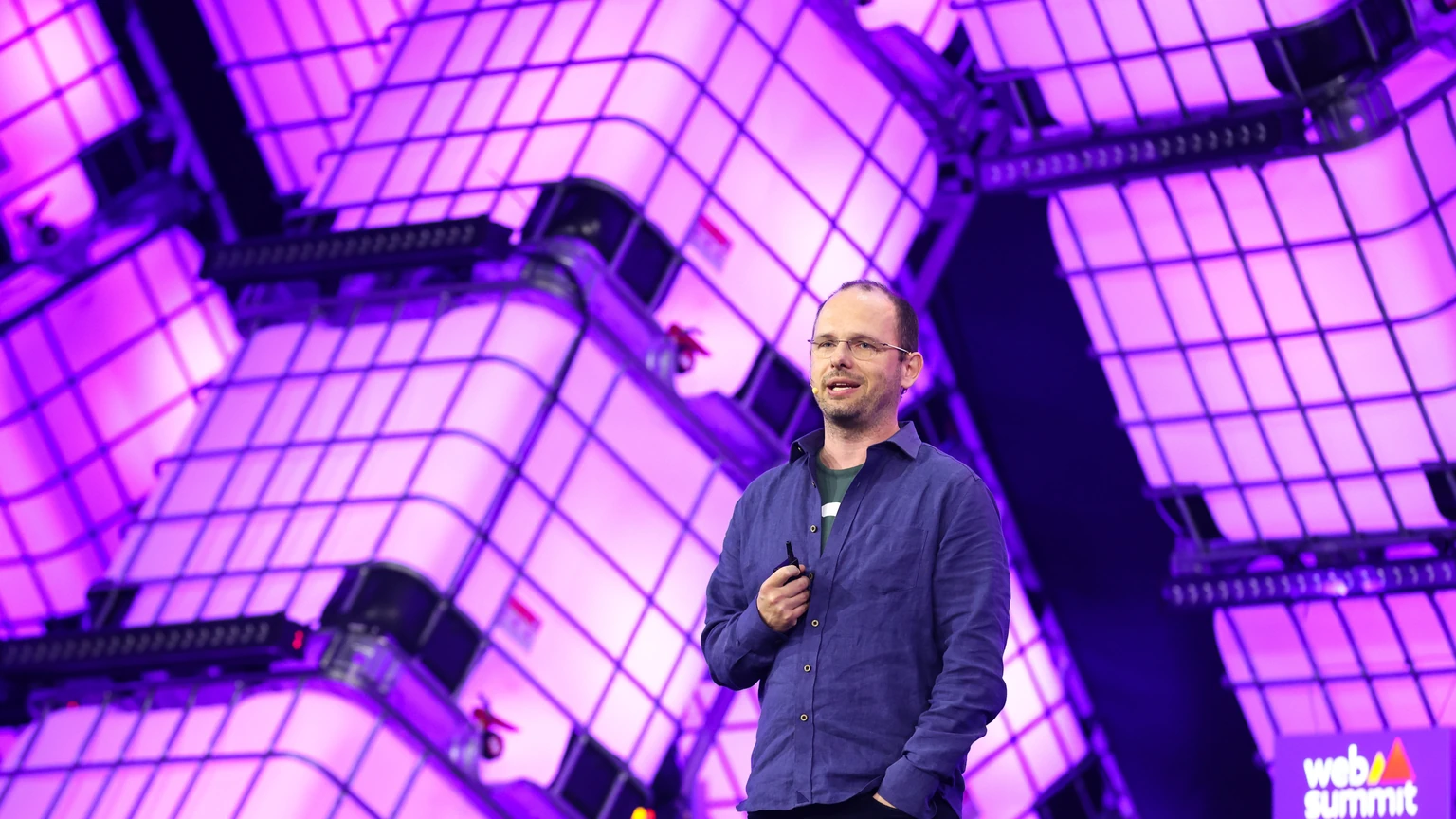Thomas Dohmke standing on a purple Web Summit stage, speaking into a handheld microphone in front of a white geometric backdrop.