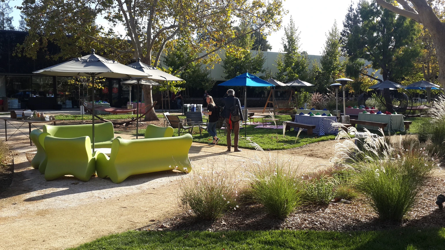 A lime-green outdoor sofa and a beige patio umbrella on a gravel path, with two people standing on a Googleplex lawn among picnic tables and trees.