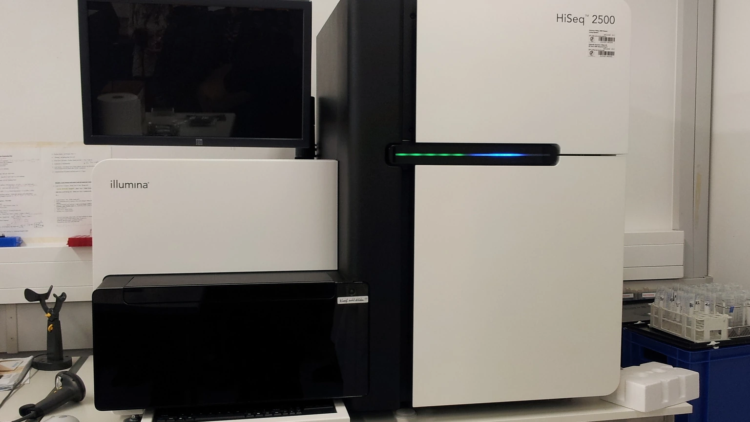 An Illumina HiSeq 2500 sequencer on a white bench, with a dark monitor, keyboard and barcode scanner to the left.
