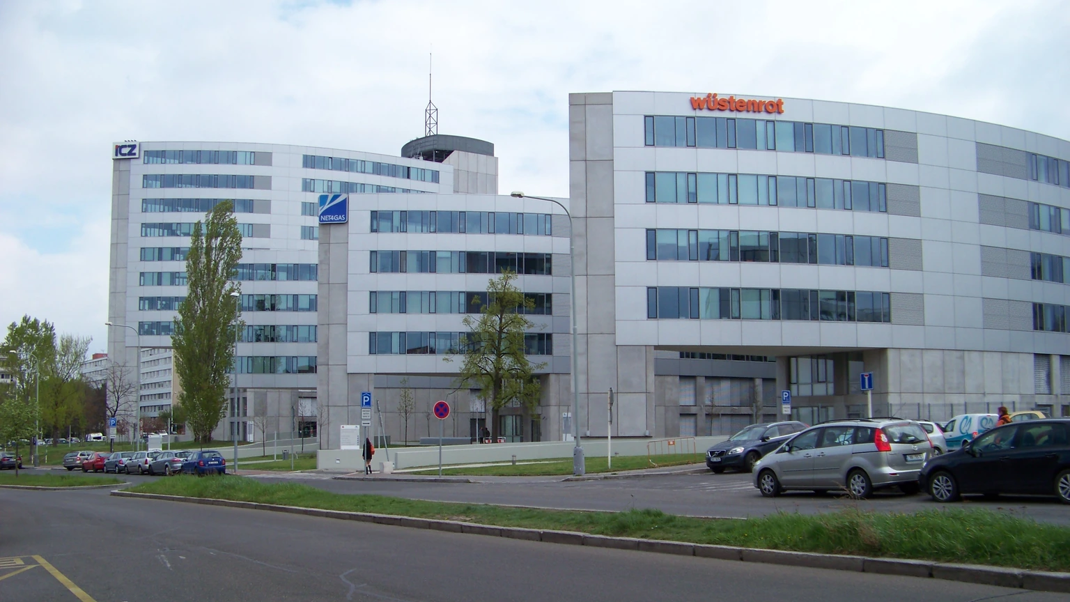 Two white curved office buildings beside an empty access road, with Wüstenrot lettering on the right roof and ICZ and Netmidas signs on the left, plus a few parked cars.