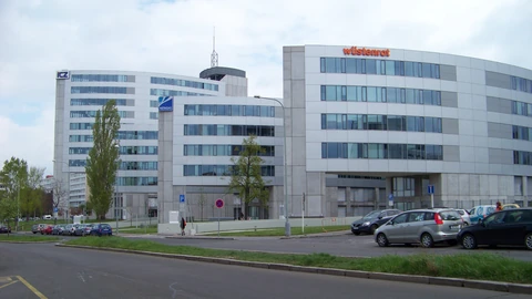 Two white curved office buildings beside an empty access road, with Wüstenrot lettering on the right roof and ICZ and Netmidas signs on the left, plus a few parked cars.