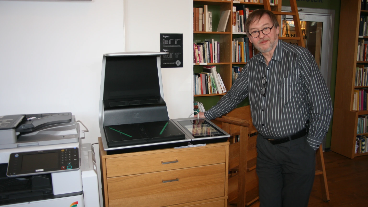 A librarian in a striped shirt standing beside a Zeutschel overhead book scanner and a multifunction copier in a library.