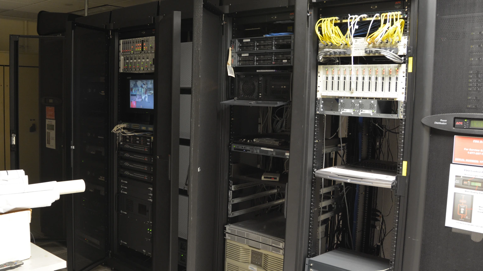 Three open black server racks with patch panels, yellow fiber jumpers and mixed network equipment in a small office server room.