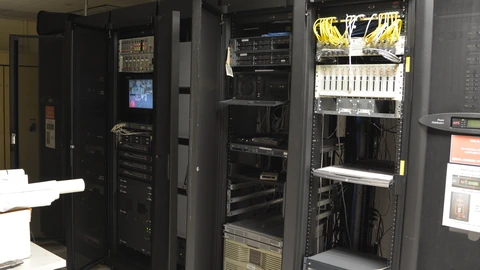 Three open black server racks with patch panels, yellow fiber jumpers and mixed network equipment in a small office server room.