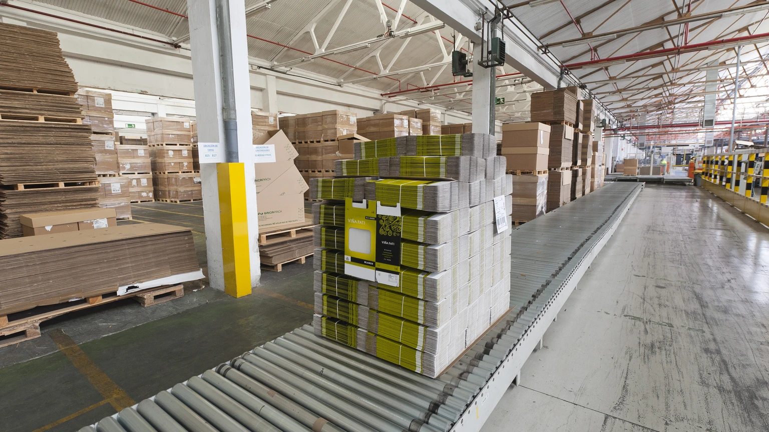 A pallet of stacked flat cardboard boxes with yellow labels sits on a roller conveyor in a warehouse, with more pallets of boxes behind it.