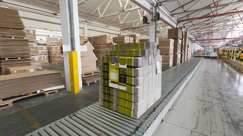 A pallet of stacked flat cardboard boxes with yellow labels sits on a roller conveyor in a warehouse, with more pallets of boxes behind it.