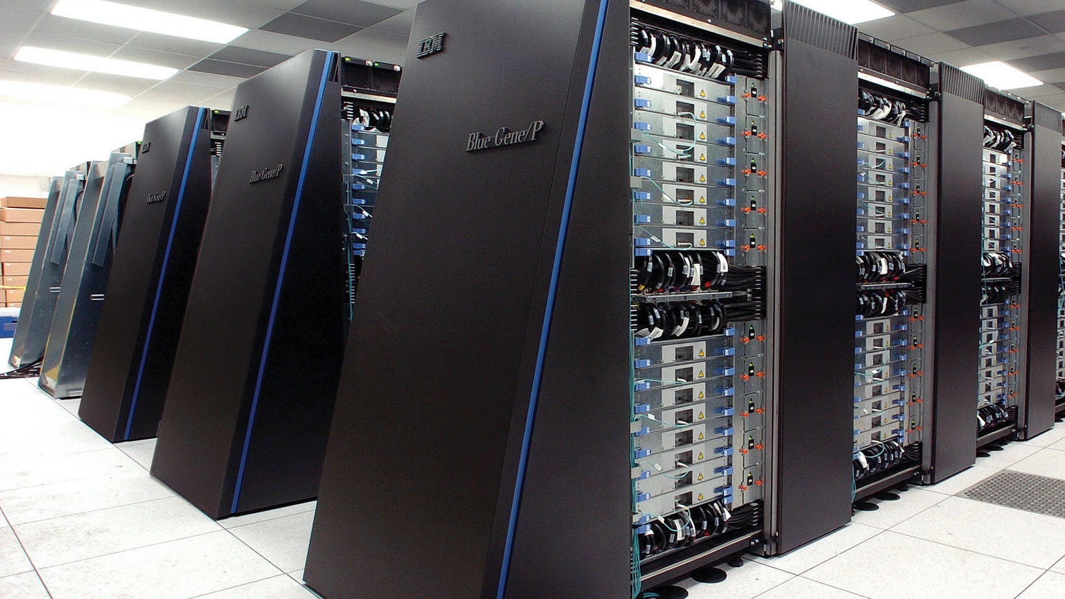 Black IBM Blue Gene/P cabinets in a white machine room, nearest door labeled BlueGene/P, cable loops visible on open racks. No people appear.