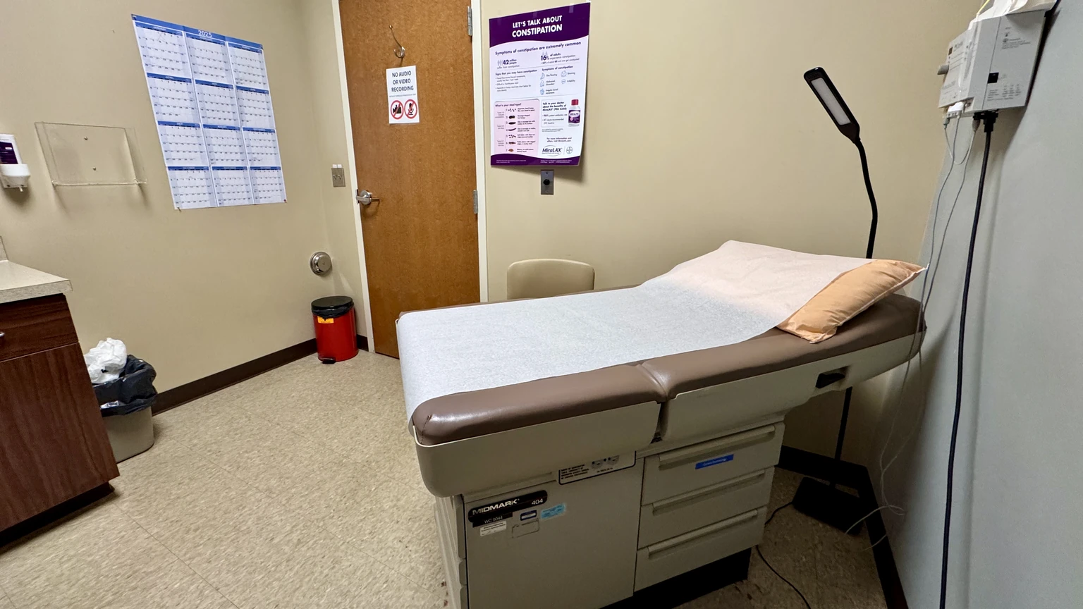 An empty primary-care examination room with a paper-covered exam table, a wall lamp, a brown door and a wall-mounted dispenser.