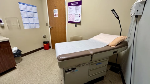 An empty primary-care examination room with a paper-covered exam table, a wall lamp, a brown door and a wall-mounted dispenser.