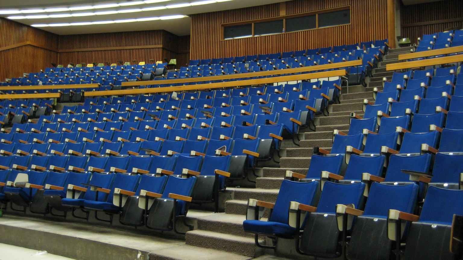 An empty lecture hall with rows of blue seats, beige steps, and a pale wooden rail, photographed from the front corner. No people appear.