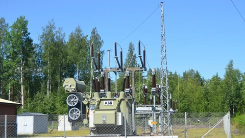 A beige ABB power transformer and insulator stack behind a chain-link fence, with birch trees and a clear sky.
