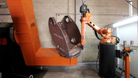 An orange ABB robotic arm on a black pedestal facing a large orange positioner that holds a brown metal housing. A cinder-block wall is behind them. No people appear.