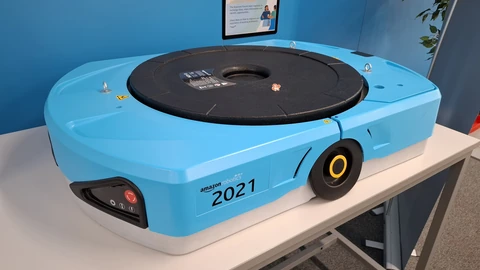 A light-blue Amazon Robotics drive unit numbered 20211 sitting on a white table against a blue wall.