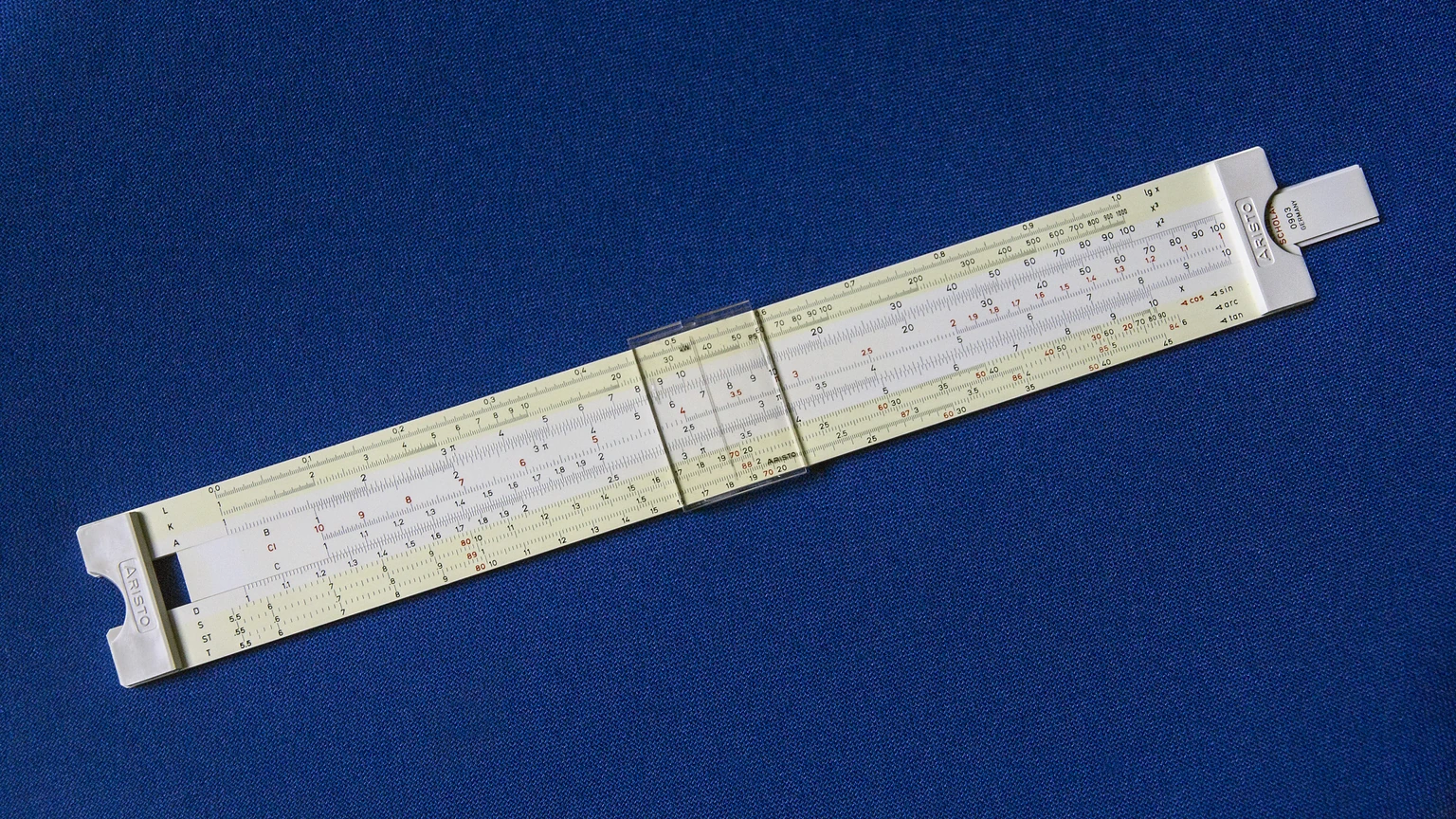 An Aristo Scholar 0903 student slide rule lying diagonally on a plain blue background, clear cursor at center, ARISTO marks on both ends. No people appear.