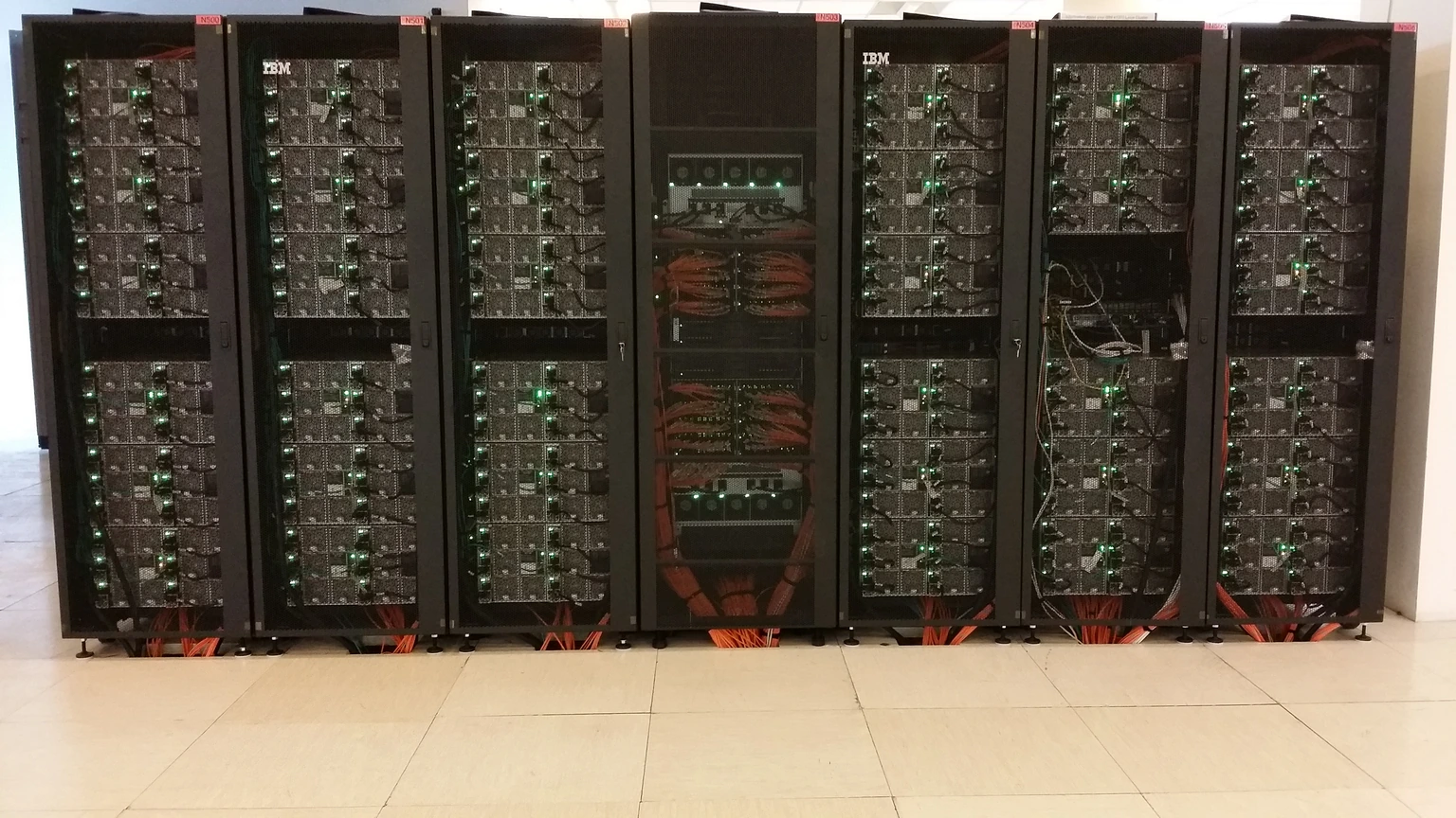 Six black IBM server racks of the Galileo supercomputer standing on a pale tiled floor, with green status LEDs and orange cables at the base.