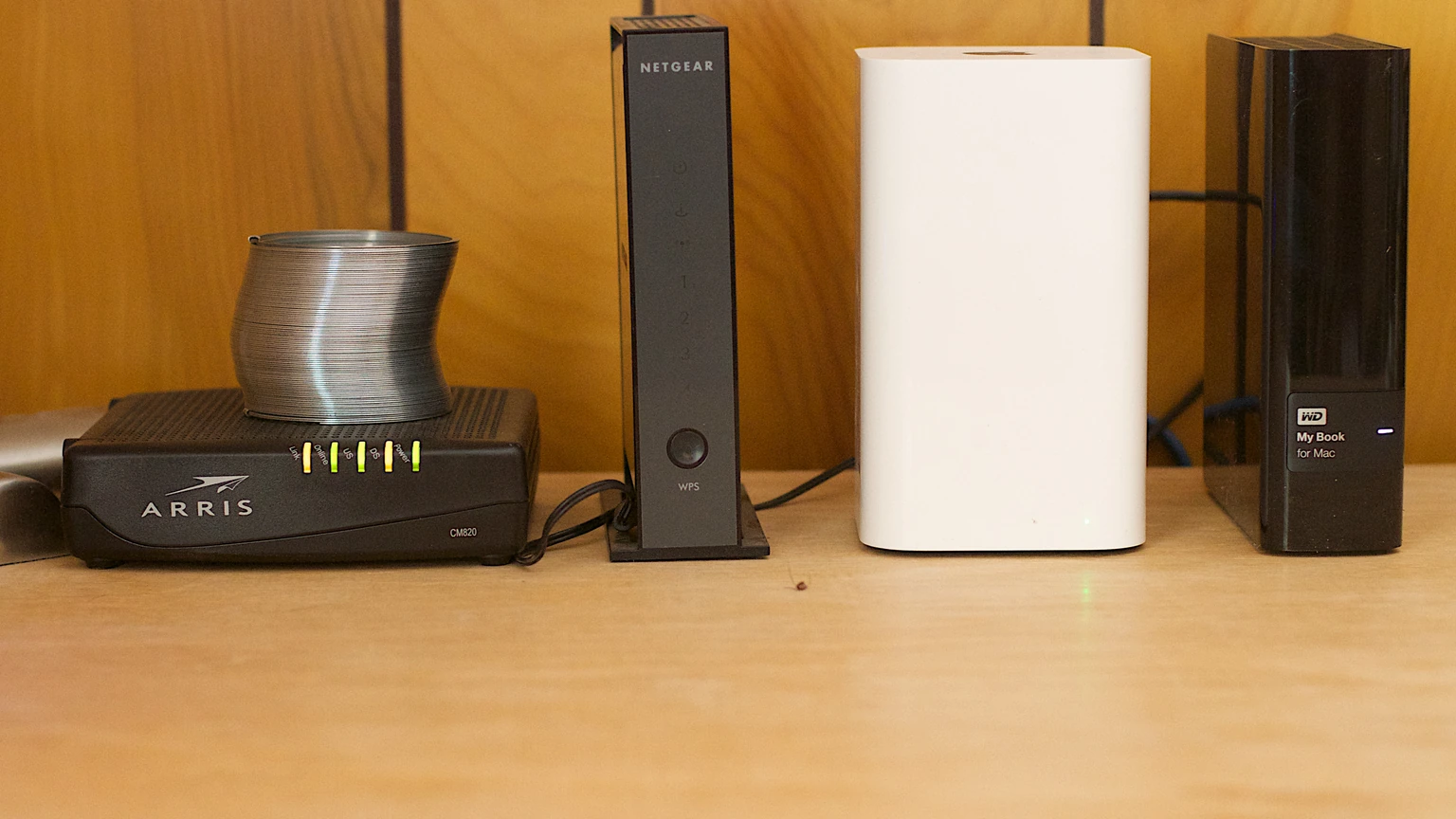 Four home-network devices on a pale desk against a wood wall: a black Arris cable modem, a tall black Netgear box, a white mesh node and a black WD My Book drive.