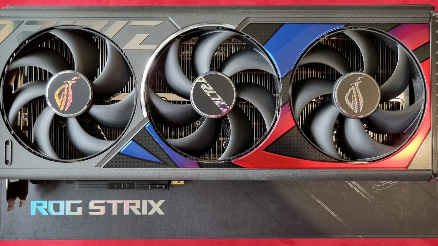 Asus ROG Strix GeForce RTX 4090 graphics card with three cooling fans on a red background, photographed from above, with no people in the frame.