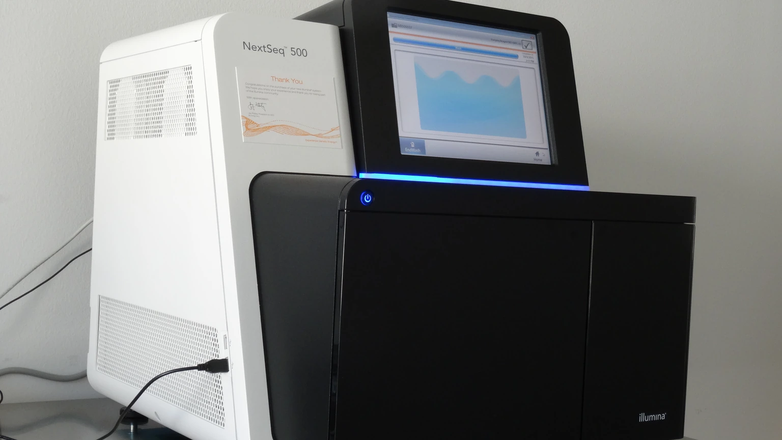 A white-and-black Illumina NextSeq 500 desktop sequencer on a bench, power cable at left, small screen showing a pale waveform, and the Illumina mark on the black door. No people appear.